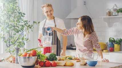 Handsome Young Man in Glasses Wearing Apron and Beautiful Girl are Making A Smoothie in the Kitchen. Happy Couple are Preparing Healthy Organic Beverage. Male and Female at Home on a Sunny Day.