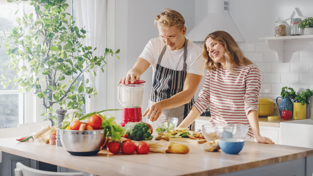 Handsome Young Man in Glasses Wearing Apron and Beautiful Girl are Making A Smoothie in the Kitchen. Happy Couple are Preparing Healthy Organic Beverage. Male and Female at Home on a Sunny Day.