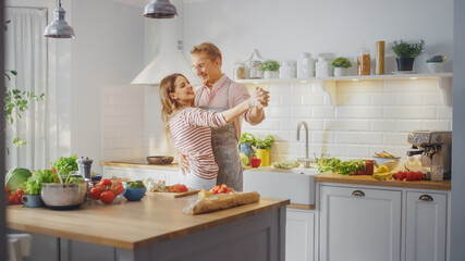 Handsome Young Man in Pink Shirt and Apron and Beautiful Girl in Striped Jumper are Creatively Dancing in the Kitchen. Sunny Modern Kitchen with Healthy Green Vegetables on Table. Happy Couple at Home