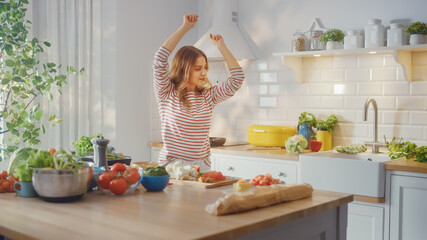 Beautiful Young Female Preparing a Healthy Organic Salad Meal in a Modern Sunny Kitchen. She is Happy and Dances to the Music. Natural Clean Diet and Healthy Way of Life Concept.