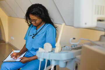 Obraz premium Young cheerful African woman operator of an ultrasound scanning machine analyzing diagnostics results of patient. Young smiling African doctor working on a modern ultrasound equipment.