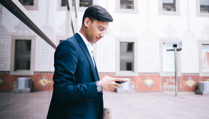 Serious hindu male millennial prosud ceo watching video online on smartphone standing outdoors, handsome businessman in trendy dressed formal outfit making banking using mobile phone application