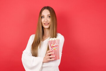 Eastern European woman holding bucket with popcorn standing over isolated red background happy face smiling with crossed arms looking at the camera. Positive person.
