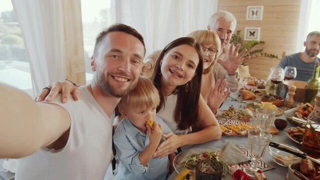 Joyous grandparents, young couple and cute little son sitting together at dinner table, smiling and looking at camera while taking selfie during holiday celebration with family at home