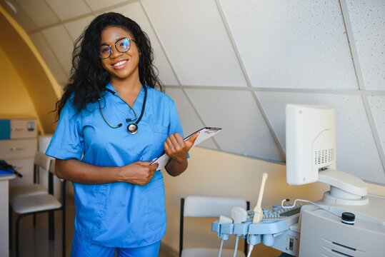 Young afro american female doctor standing in clinic office near modern ultrasound machine.