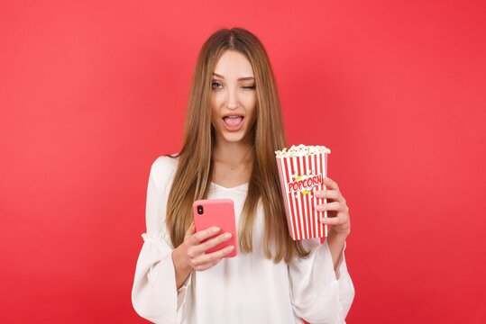 Eastern European woman holding bucket with popcorn standing over isolated red background,taking a selfie  celebrating success