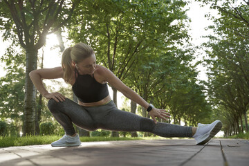 Fototapeta premium Warming up outdoors. Full length of beautiful fitness woman in sportswear stretching her legs while exercising in city park on a sunny summer morning