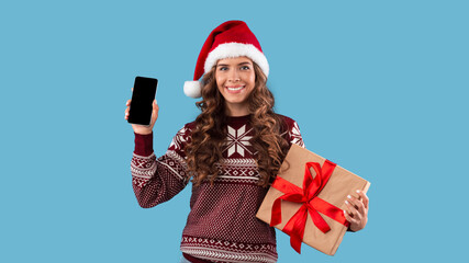 Smiling girl in Christmas outfit holding gift box and cellphone with blank screen over blue background, space for design