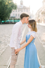Fototapeta premium Handsome young man kissing his charming pretty girlfriend on forehead in the street in the sunny morning. Romantic couple in the street in ancient European city