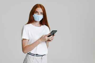 Image of displeased ginger girl in face mask using mobile phone