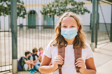 Blond teen girl standing outside school, wearing a protective mask. A group of kids behind, sunny day. Concept back to school new rules during coronavirus pandemic, Covid-19.
