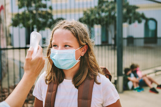 Hand holding the non-contact infrared thermometer to measure girl's forehead outside school. She wears a protective face mask. Concept back to school, coronavirus and social distance.