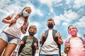 Low angle view of Children standing wearing protective face masks and backpacks. They are in a row looking at the camera. It's daytime with cloudy blue sky behind. Back to school after coronavirus.