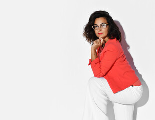 Self-confident woman with long curly hair in eyeglasses and formal suit red jacket and blue trousers stands side to camera