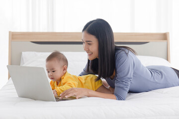 An Asian mom is working on a laptop in bed and with her baby next to the concept of work at home.