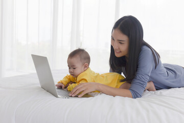 An Asian mom is working on a laptop in bed and with her baby next to the concept of work at home.