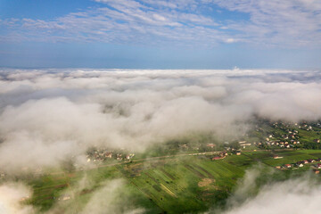 Aerial view of white clouds above a town or village with rows of buildings and curvy streets between green fields in summer. Countryside landscape from above.