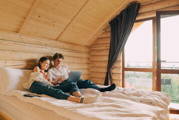 Happy couple lying on bed in hotel with wooden interior and watching movies on laptop with happy face. Positive man and woman using laptop on the bed in the apartment.