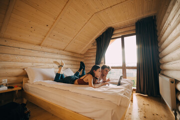 Beautiful young couple relaxing in an apartment with wooden interior, lying on the bed and using a laptop. A woman and a young man watching a movie on the bed in a hotel room