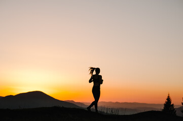 Silhouette against sky at sunrise while running. Runner fitness girl in sport tight clothes. Athlete woman on the road in the mountains. Bright sunset and blurry background. Workout wellness concept.