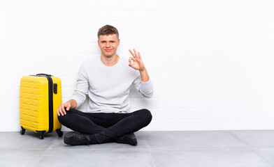 Young handsome man sitting on the floor with a suitcase showing an ok sign with fingers