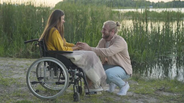 Side view of happy Caucasian man giving engagement ring to handicapped woman on river bank at sunset. Portrait of romantic boyfriend asking disabled girlfriend to marry him.