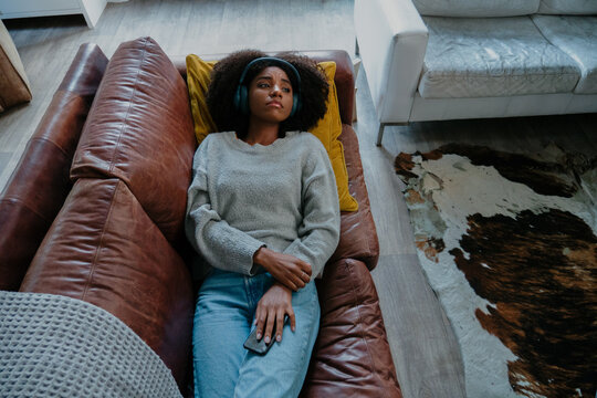 melancholic mixed race female wearing headphones listening to sad moving while holding smartphone lying on leather sofa in modern lounge.