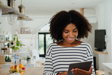 Beautiful smiling ethnic female scrolling on digital tablet standing in modern kitchen. 