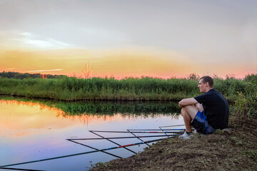 Calm relaxed man fishes on a lot of fishing while sitting on the shore of the lake at sunset