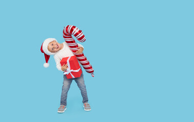 Portrait of a satisfied little child boy in christmas Santa hat. laughing isolated over blue background. Holds a gift box and big candy cane. Preparing for the New Year holidays