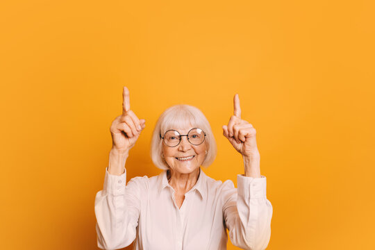 Portrait of smiling old woman with short gray hair and round glasses wearing white blouse, pointing fingers up. Senior woman isolated over orange background.