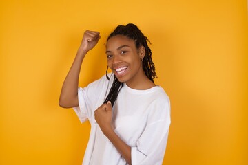 Profile photo of excited Young dark skinned woman with braids hair wearing casual clothes trendy isolated over yellow background raising fists celebrating black friday shopping