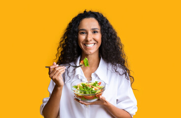 Healthy Eating. Smiling curly woman enjoying fresh vegetable salad over yellow background
