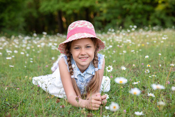 Cute little girl in summer dress and hat walks in the garden among daisies
