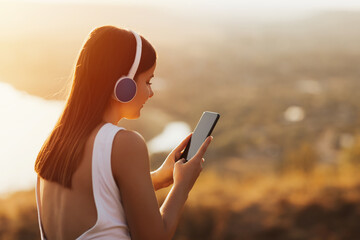 Pretty woman looking at phone at sunset. Close up of joyful young woman with headphones relaxing in the park and smiling. Phone with black screen. © eduard