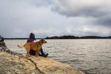 Young woman girl with dog on rocky coast looking at the sea. Autumn Fall Concept. View of Scandinavian Stockholm Archipelago on cloudy sky background. Pine forest islands on the horizon.