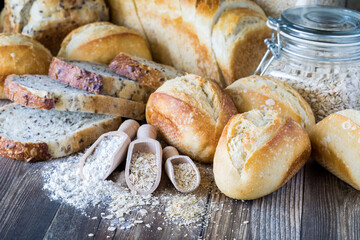 Various buns and loaves of bread on a wooden table with wooden scoops of ingredients used to make bread in front.