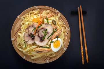Noodles with pieces of meat and an egg in a bamboo plate on a dark table. Asian food concept. View from above