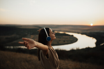 Young beautiful woman listens to music in summer during sunset. Smiling pretty woman with closed eyes and hands up feeling positive emotions. Copy space.