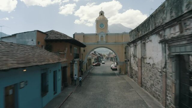 Drone aerial flying low between houses to the historical Santa Catalina Arch with volcano in the background in Antigua guatemala