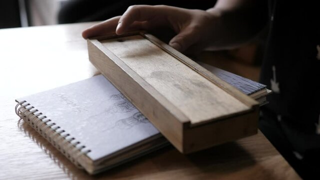 young woman closes wooden vintage pencil box on paper notebook with sketch sitting at table in light room closeup slow motion