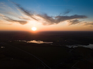 Fototapeta na wymiar Aerial view of the sunrise over dark rural landscape with lake and river. Rising sun at blue cloudy sky over horizon and dark surface with reflecting water.