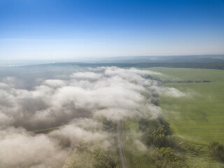 Aerial view on white clouds over rural landscape with fields, forest, meadows and road under clear blue sky. Summer sunny day.