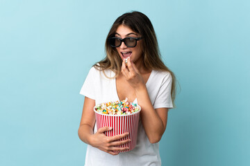 Young caucasian woman isolated on blue background with 3d glasses and holding a big bucket of popcorns while looking side