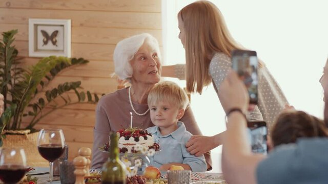 Joyous senior woman sitting at dinner table while young granddaughter bringing birthday cake and kissing her cheek; family taking pictures with smartphones and clapping hands during home celebration