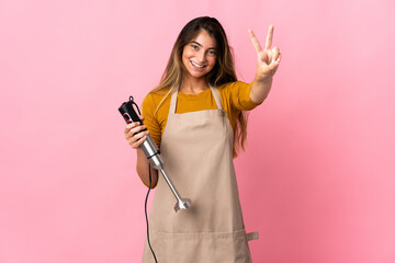 Young chef woman using hand blender isolated on pink background smiling and showing victory sign