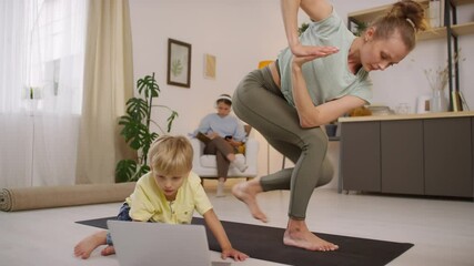 Tracking of young woman in sportswear doing yoga and stretching on mat while her little son using laptop and teenage daughter listening to music and doing homework in background