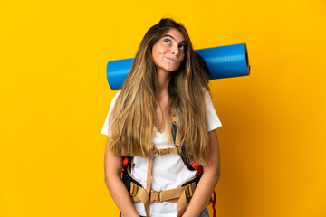 Young mountaineer woman with a big backpack isolated on yellow background and looking up