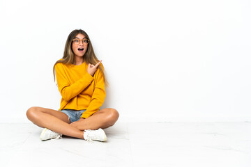 Young woman sitting on the floor isolated on white background surprised and pointing side