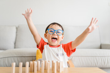 Happy smart and nerd toddler asian boy raised hand when he win play wooden toy block at home.Boy child kid wearing glasses.myopia or Short sighted.Positive human emotion, Attitude and self esteem.